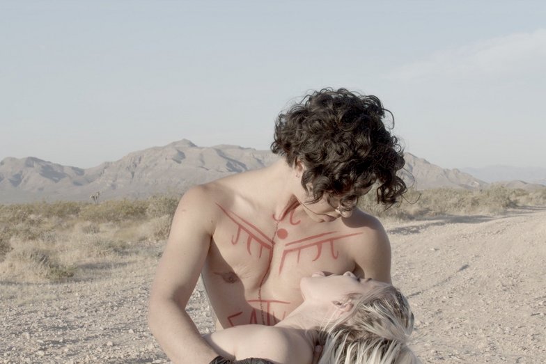 One person holds another person's torso on their lap. The chest is painted with red symbols. The person on the lap licks the other person's chest.