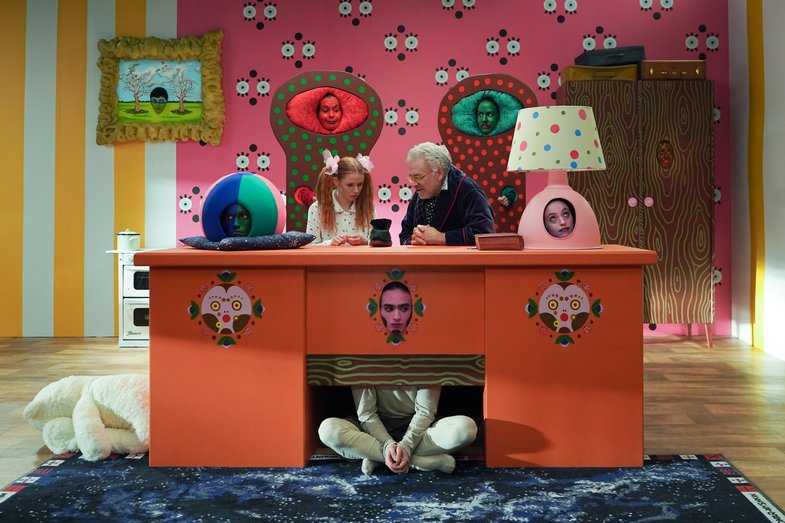 Two people sit behind an oversized desk and talk. The backdrop is dreamlike. The pieces of furniture have human faces.