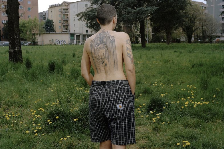 Photograph of a person in a meadow in a housing estate. They wear only pants, the back is tattooed.