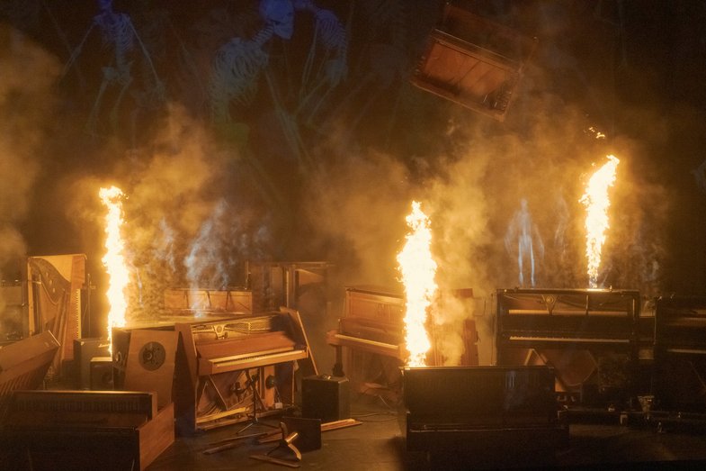 Foto von Pianos auf einer Bühne, aus einigen sprüht eine Fontäne aus Feuer.